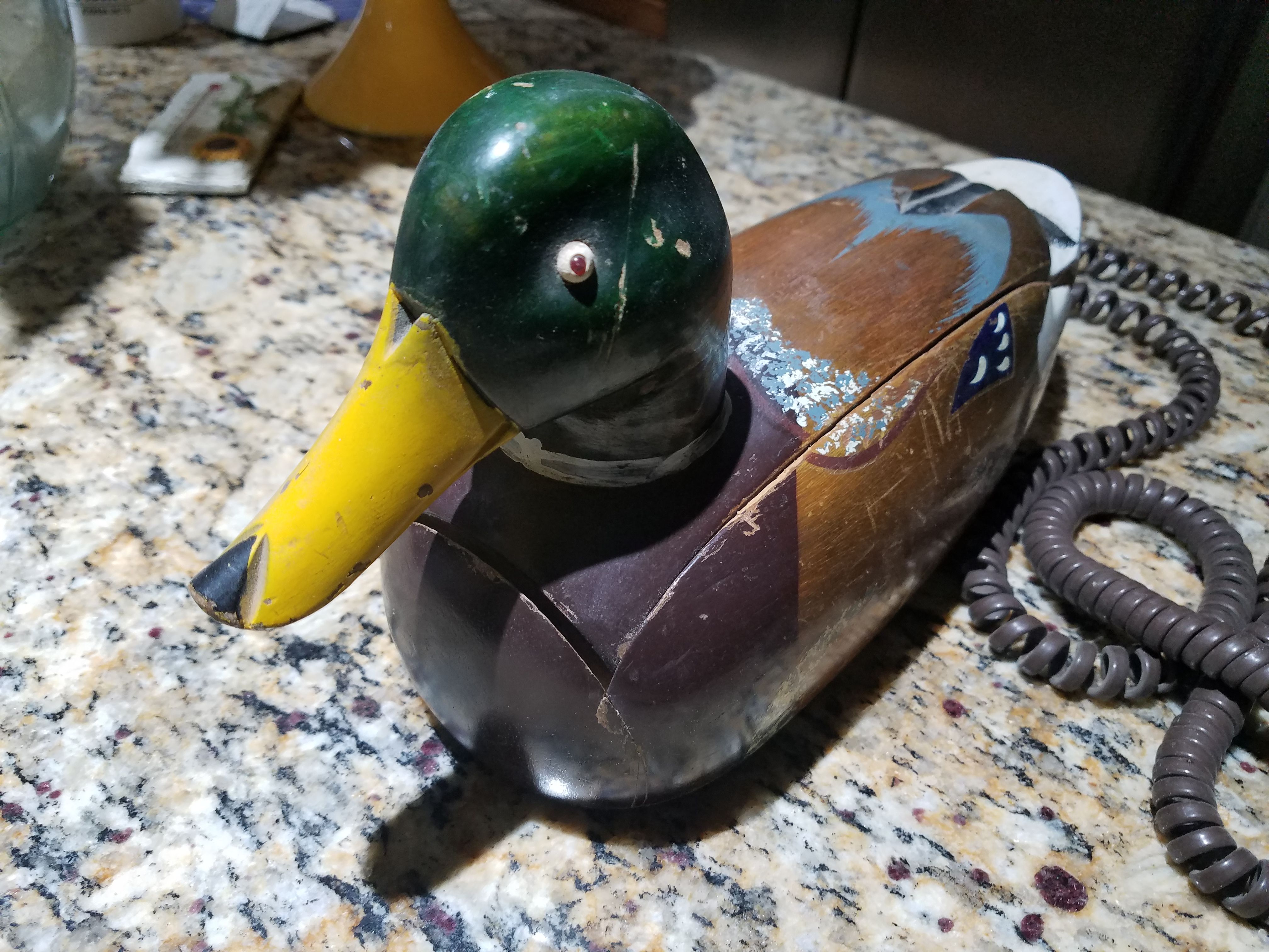 Image is of a corded phone in the shape of a duck. It's quite worn and appears to be made of wood. Its beady little eye, likely a literal bead, looks at the viewer reproachfully.